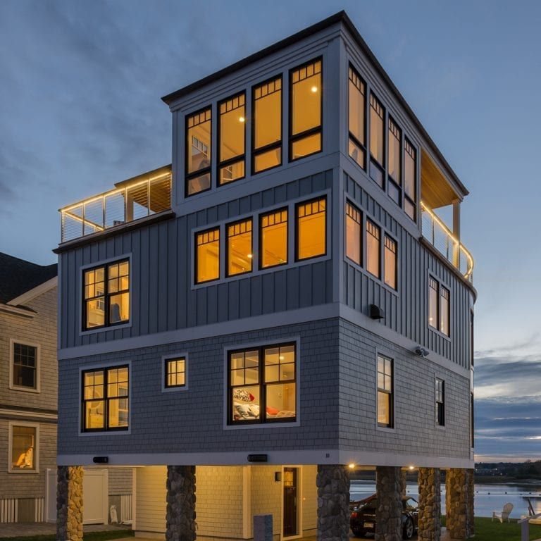 Modern three-story house with blue siding, large windows illuminated at dusk, and a rooftop deck in Wells, Maine, set against a twilight sky.