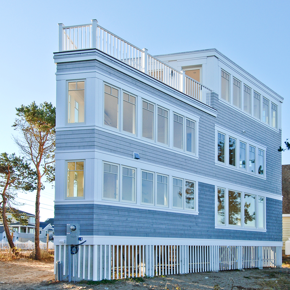 A modern three-story custom home with large windows and a rooftop deck, painted in light blue, enclosed by a white picket fence.