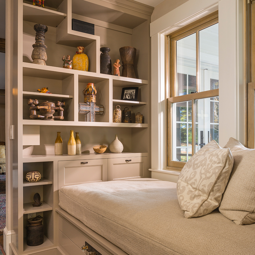 Cozy reading nook with built-in seating and storage, filled with various decorative items and books, recently renovated to enhance the comfort of any Maine home, next to a large window.