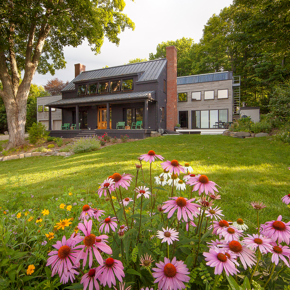 Modern two-story home with large windows and a mix of wood and dark paneling, surrounded by a lush garden with pink and yellow flowers in the foreground, located in Wells, Maine.