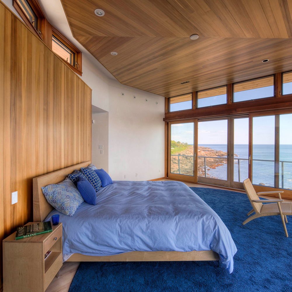 Modern bedroom in a Maine custom home, with wooden walls and ceiling, featuring a large bed and expansive windows overlooking a sea view.