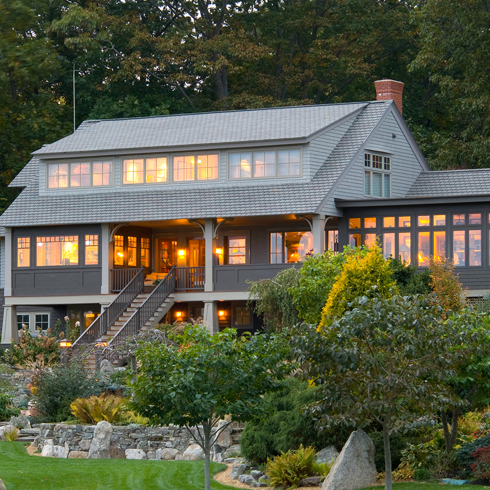 Large two-story custom home with a grey exterior and multiple windows lit up at dusk, surrounded by lush green landscaping.