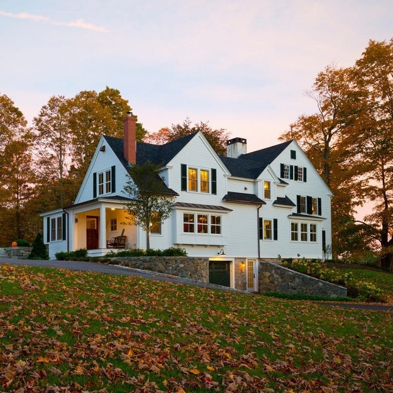 A large custom white house with black trim at dusk, featuring a covered porch and autumn leaves on the ground.