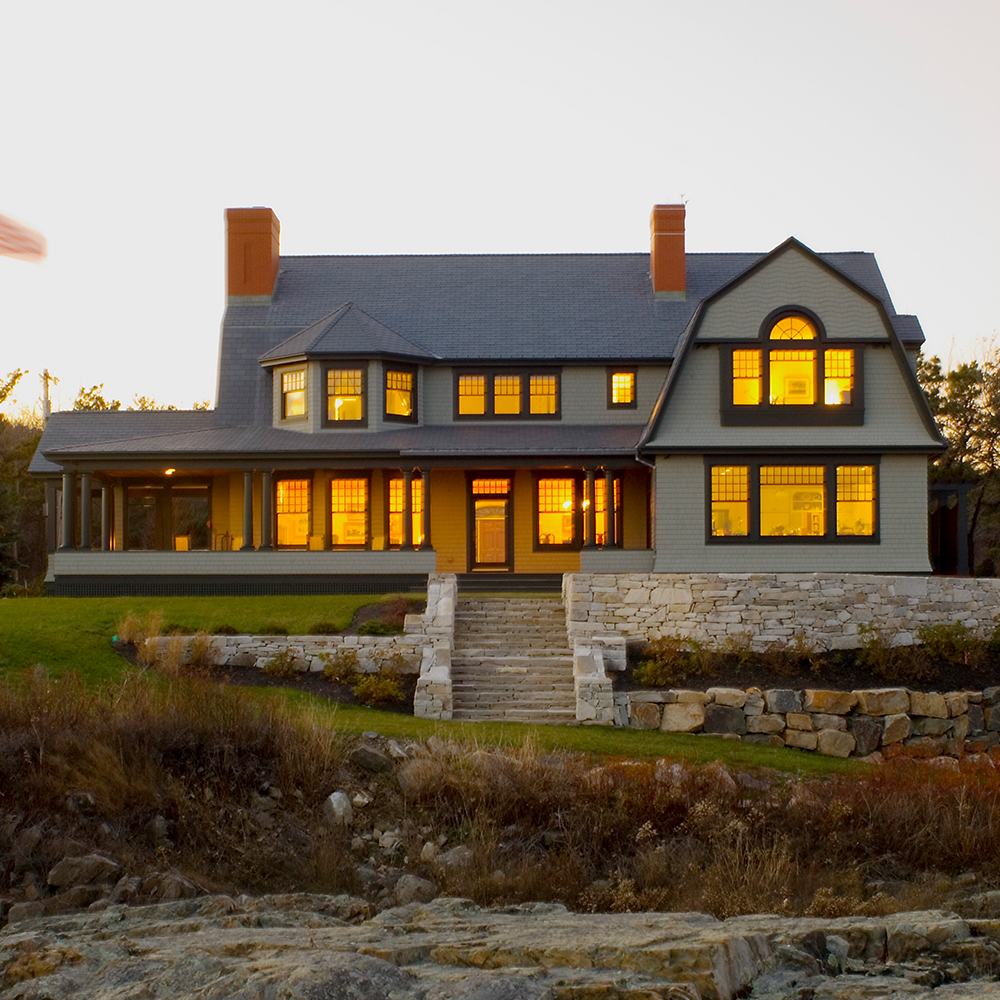 A large two-story home with lit windows at twilight, featuring a stone pathway and surrounding natural landscape.
