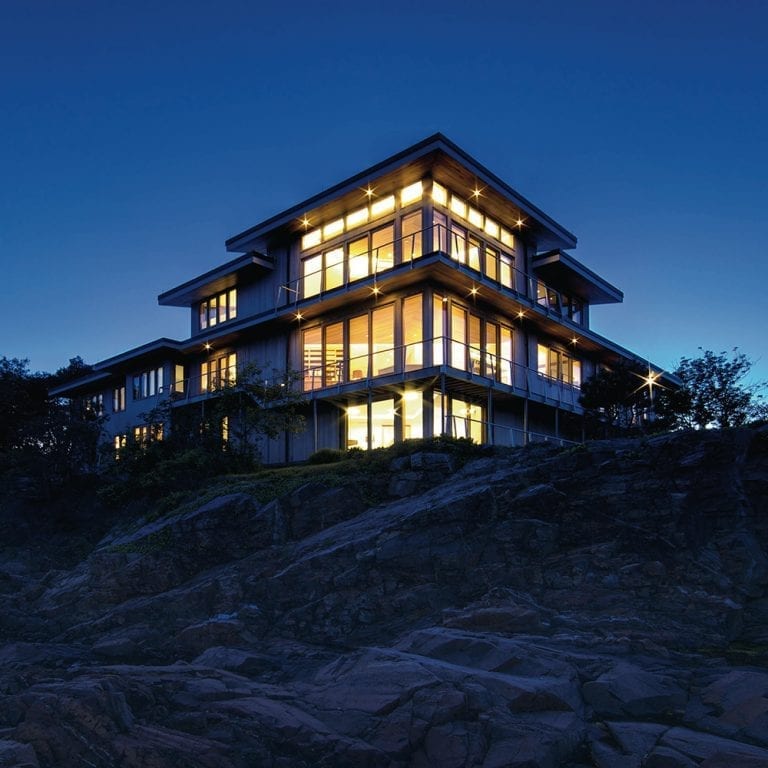 Modern multi-story custom home illuminated at twilight, perched atop a rugged hill with dense trees in the background.