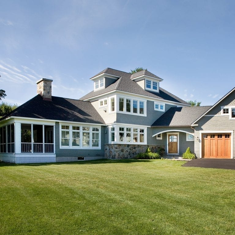 Large suburban custom home with white siding, stone accents, and a detached garage, surrounded by a well-manicured lawn under a clear sky.
