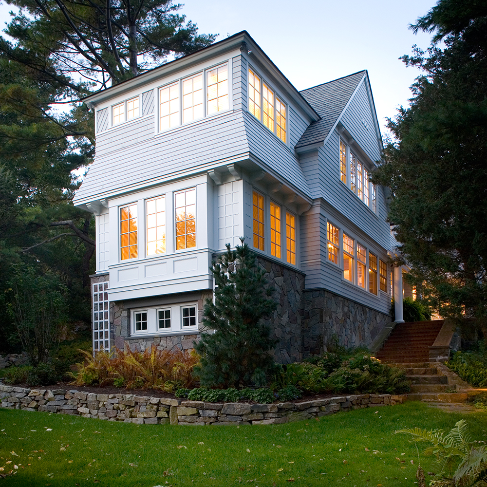 A large two-story custom home in Wells, Maine with white siding and stone foundation, illuminated windows, surrounded by lush greenery at dusk.