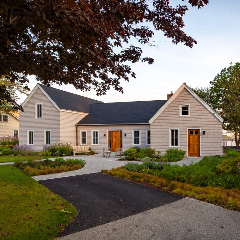 A modern single-story custom home with a light gray exterior and a neat garden pathway, surrounded by lush trees at dusk.