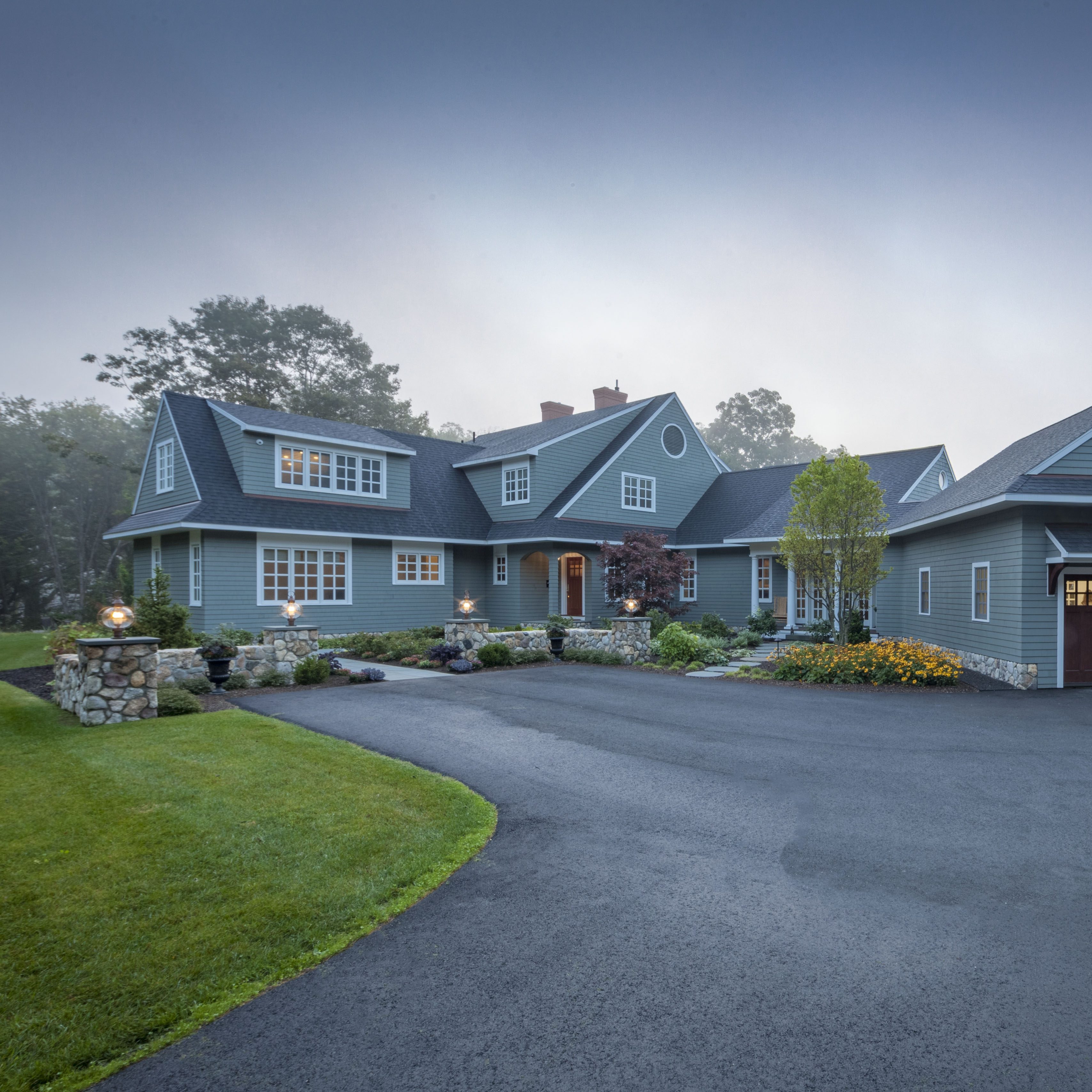 A large, two-story blue home with white trim and a driveway, surrounded by lush landscaping, during a misty evening.