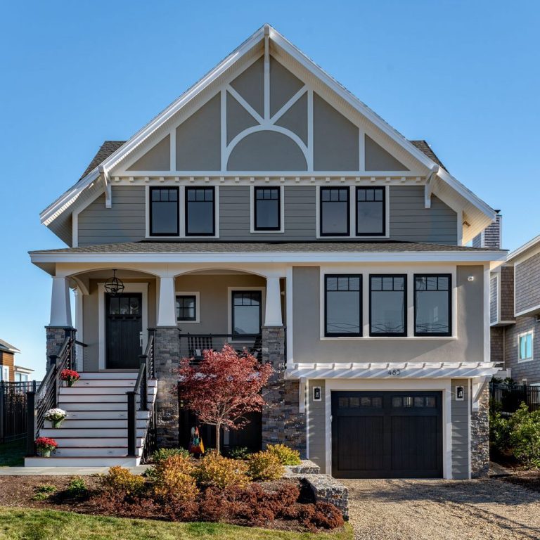 Modern three-story house in Wells, Maine, with gray siding, white trim, black windows, and a garage, surrounded by a manicured lawn and a small garden.