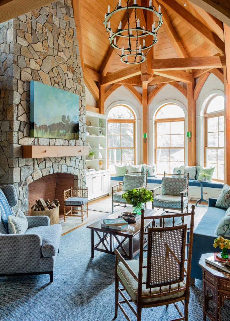 A cozy, well-lit living room in Wells, Maine, with a stone fireplace, wooden beams, arched windows, and elegant furniture arranged on a blue rug.