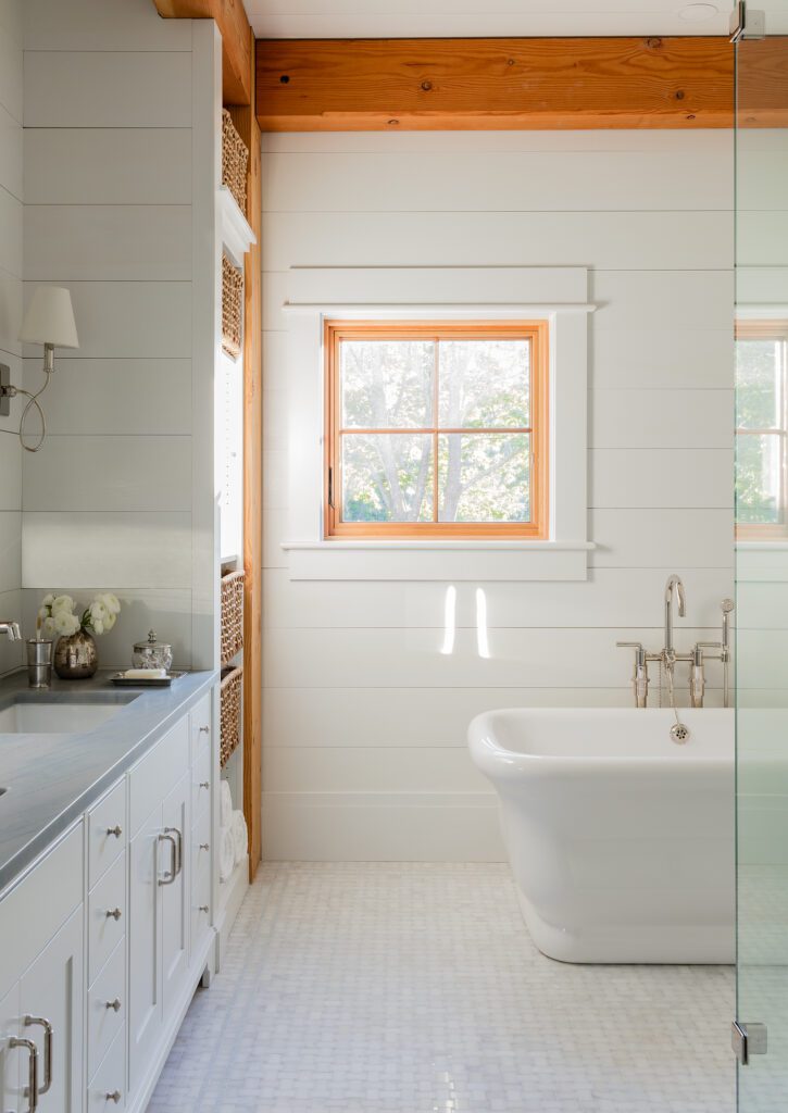 A bright, modern bathroom with white shiplap walls, a standalone tub, wooden ceiling beams, and a window showing greenery outside showcases recent renovations.