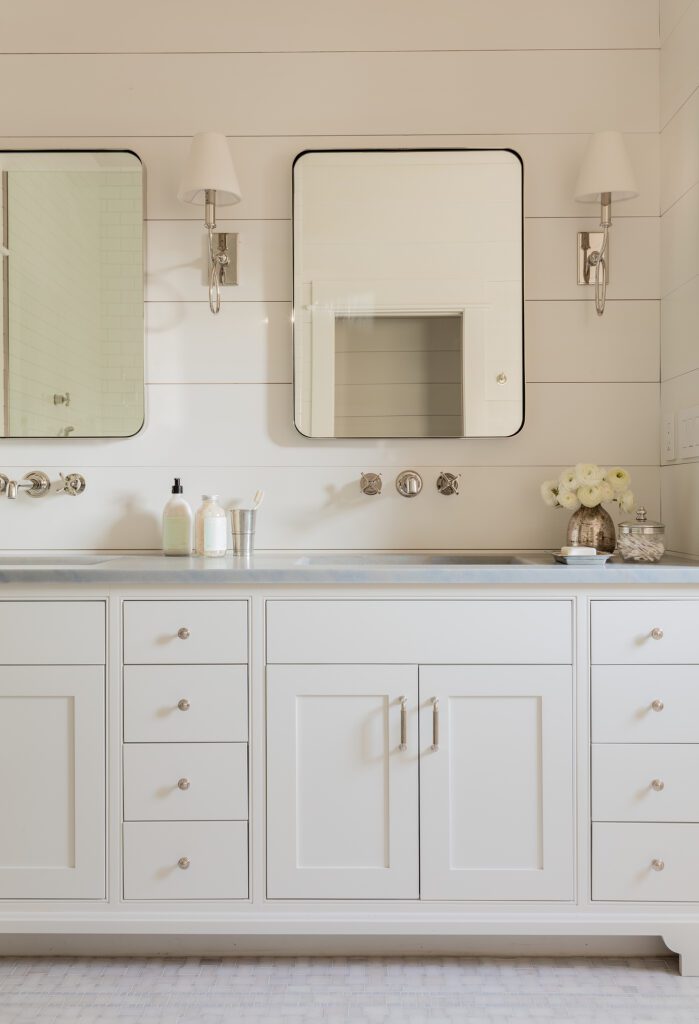 Elegant bathroom vanity with dual sinks, white cabinetry, and two mirrors flanked by wall-mounted lamps. Fresh flowers and bath products are on the countertop in this custom home.