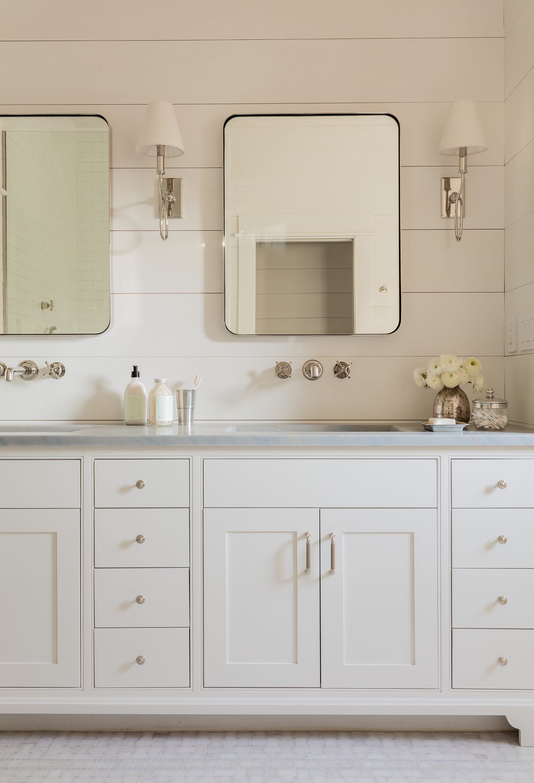 Elegant bathroom vanity with dual sinks, white cabinetry, and two mirrors flanked by wall-mounted lamps. Fresh flowers and bath products are on the countertop in this custom home.