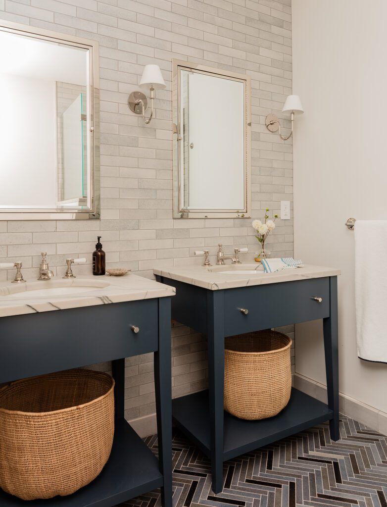 A modern bathroom with a blue double vanity, two mirrors, sconce lighting, and a chevron tile floor. Wicker baskets under the sinks provide storage in custom homes.