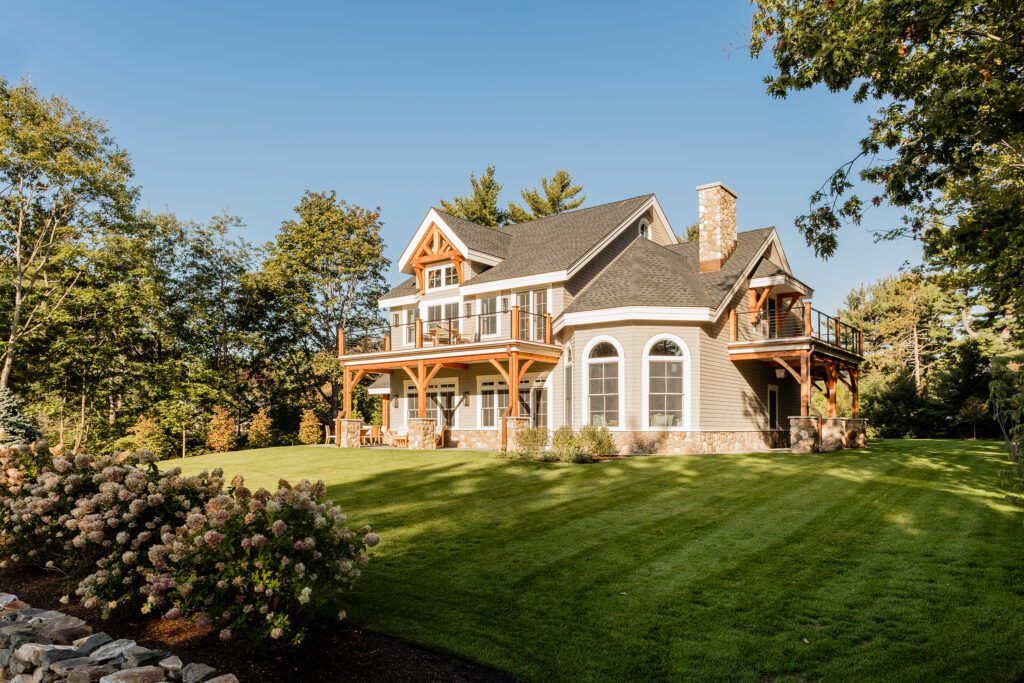 Large two-story house with a wraparound porch and dormer windows, surrounded by landscaped lawn and trees under a clear blue sky, perfect for custom homes.