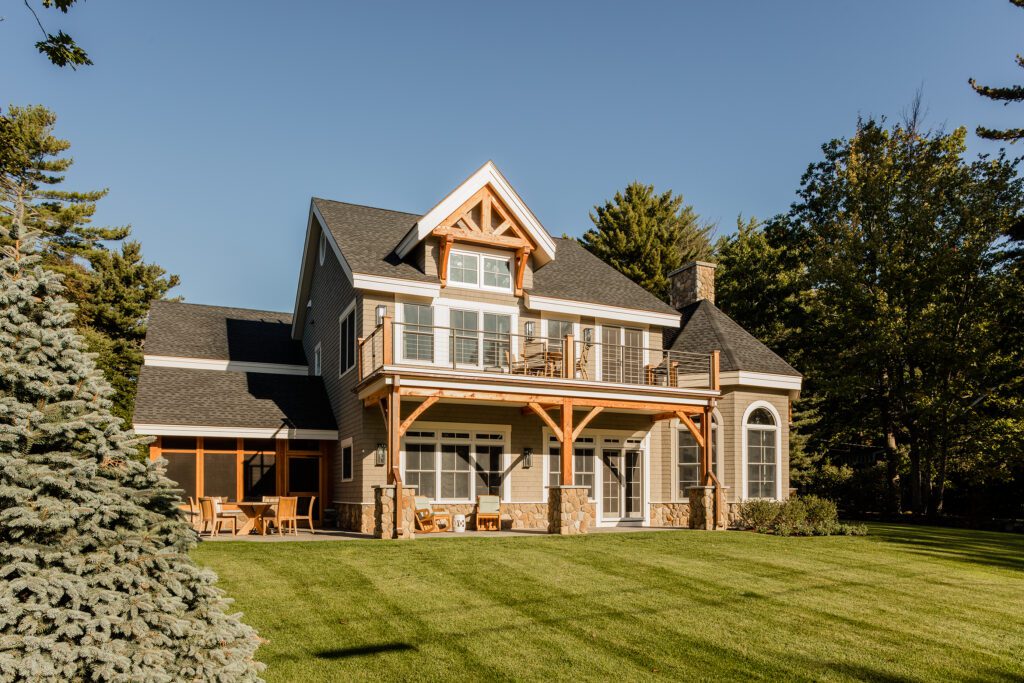 A luxurious two-story home in Maine with a large wooden deck, expansive windows, and a well-manicured lawn, surrounded by lush green trees under a clear blue sky.