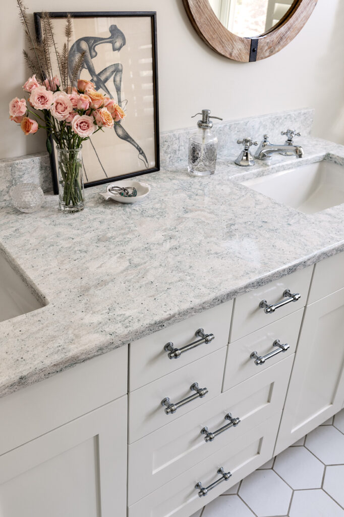 Modern bathroom vanity with white cabinets, granite countertop, dual faucets, and a framed abstract art piece above. The decor includes a vase of pink roses and is part of a home remodeling project by