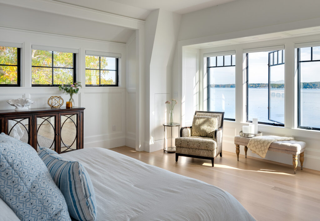 Bright, modern bedroom with large windows overlooking a lake in Maine, featuring elegant furniture and hardwood floors.