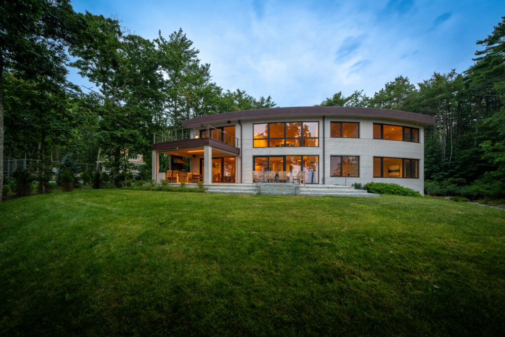 Modern home in Wells, Maine, with large windows and curved architecture surrounded by trees at dusk, lights inside are on, two people visible.