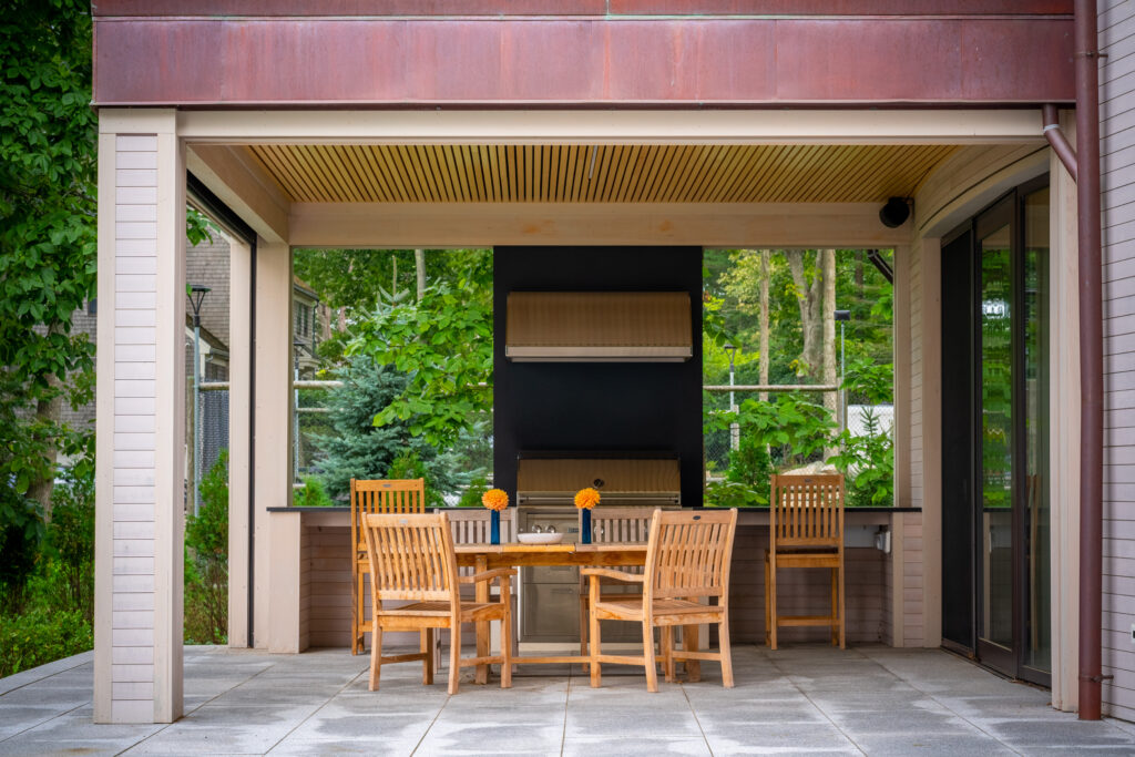 Modern outdoor dining area with a wooden table and chairs set under a covered patio, surrounded by lush greenery in Wells, Maine.
