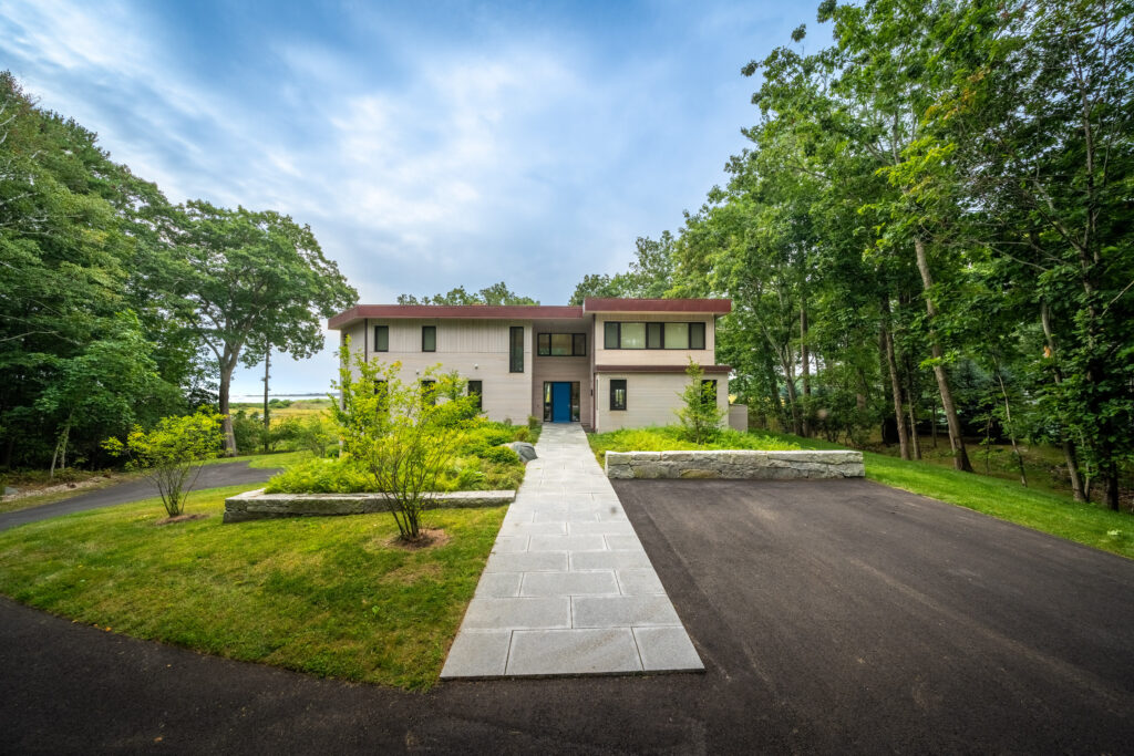 Modern two-story house with a green and beige exterior, surrounded by lush trees, featuring a stone walkway and recent renovations.