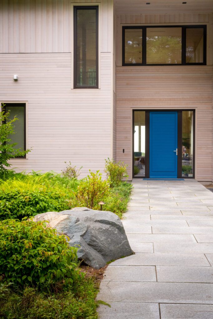 A modern house exterior in Maine with pale pink siding, featuring large windows and a bold blue door, complemented by a stone pathway and lush greenery.