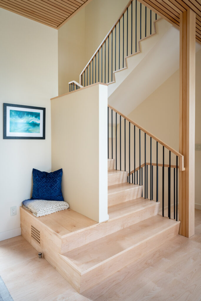 Interior view of a modern staircase in a Maine custom home, featuring wooden steps, black railings, and a built-in seating area with a blue cushion and a framed ocean painting on the wall.