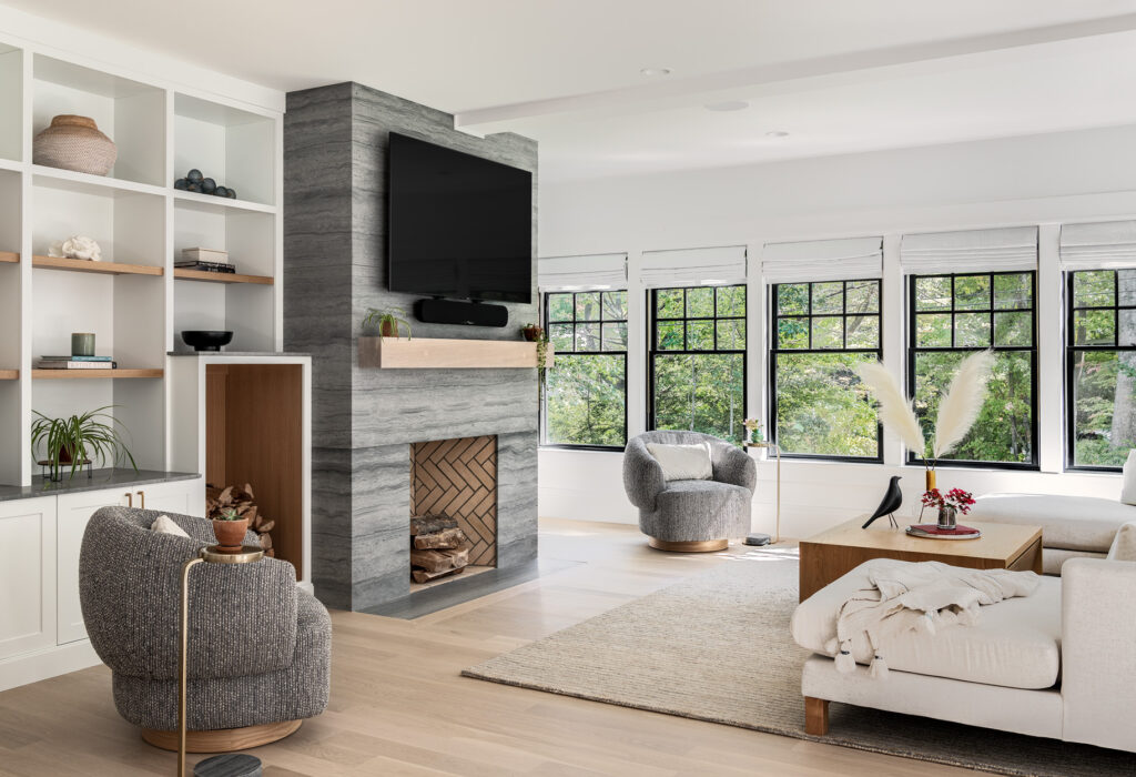 Modern living room with neutral colors, featuring a fireplace, a large tv, comfortable seating, and floor-to-ceiling windows surrounded by greenery in custom homes.
