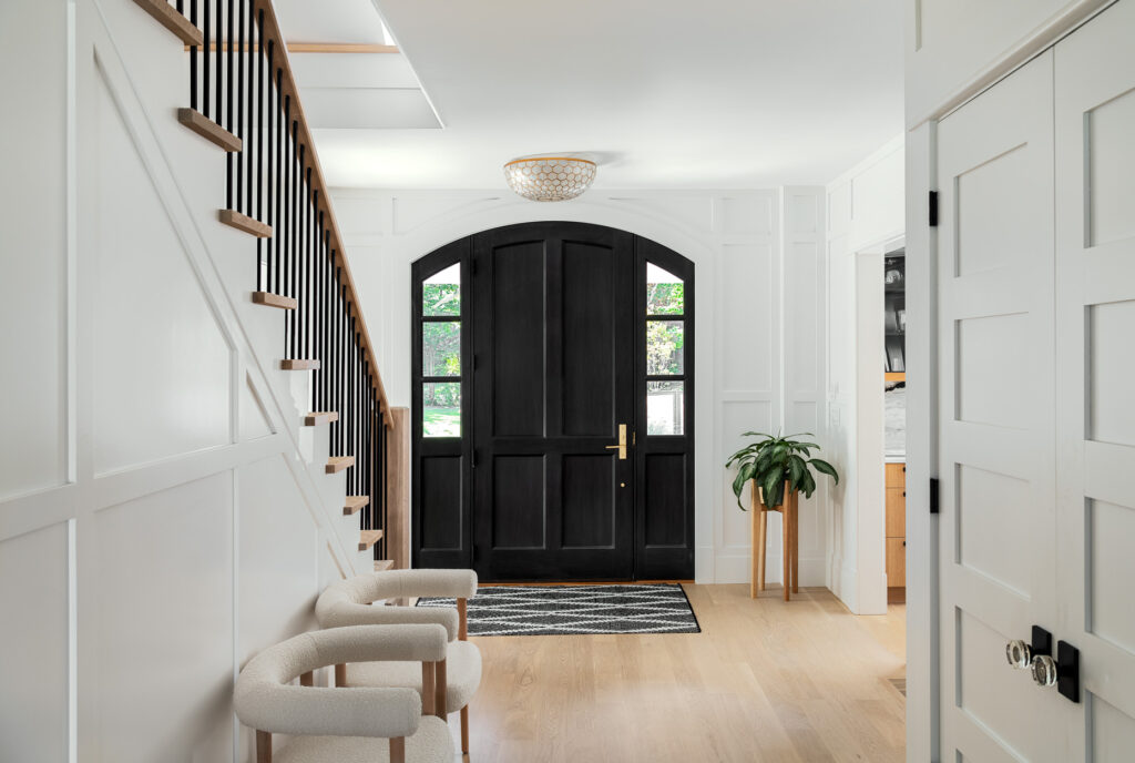 Elegant home entryway with a black arched door, white walls with wainscoting, wooden staircase, and beige upholstered chairs, perfect for custom homes.