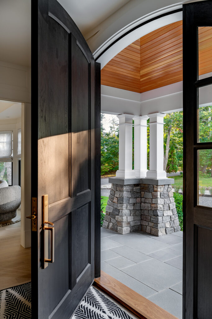 Open door leading to a custom home's covered patio with stone pillars, wooden ceiling, and a view of a garden.